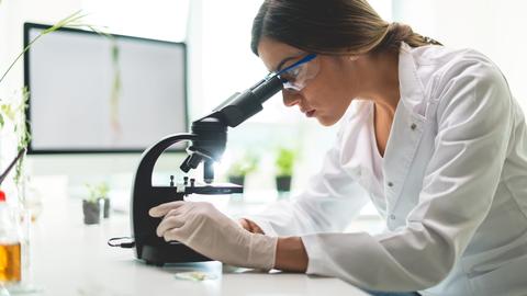 Young woman in white wearing gloves leans over microscope.