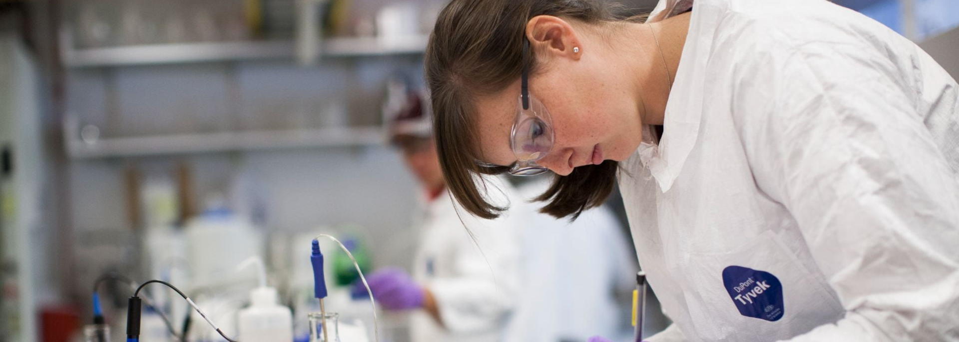 A student in a laboratory wearing a white coat and gloves writes in a notebook.